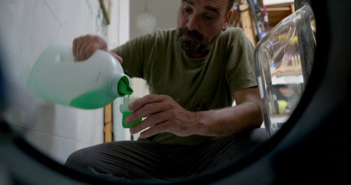 This shot was taken with the camera inside the washing machine. A man is pouring washing-up liquid: A bearded man pours washing-up liquid from a large bottle into a smaller cup to put it into the wash