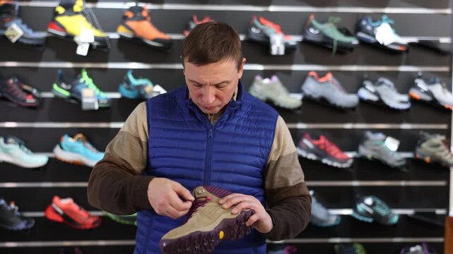 Man chooses specialized shoes in store, near showcase with tracking cross-shoes, buyers think about advantages and disadvantages of winter trecking boots. 