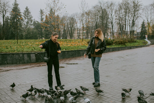 Female friends feeding pigeons on rainy autumn day in city square