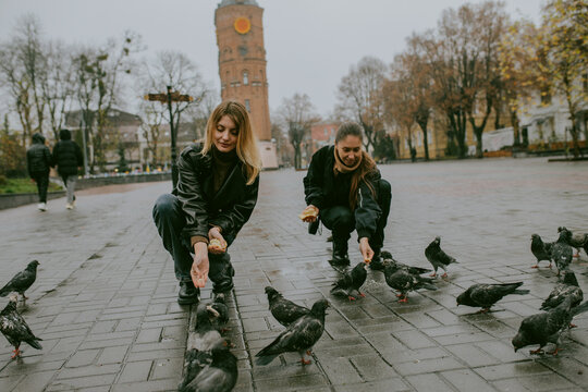 Female friends feeding pigeons on rainy autumn day in city square