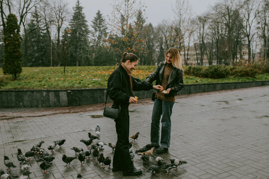 Female friends feeding pigeons on rainy autumn day in city square