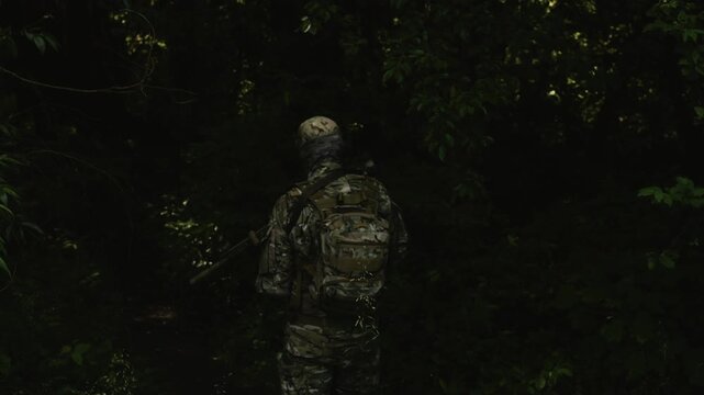 Camouflaged soldier in the middle of a dark forest on a recon mission. Walking and aiming a AR-15 carbine rifle with a red collimator and a magnifier
