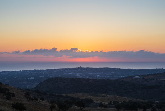 Scenic sunset over Analipsi coastal town, viewed from the hiking trail of Mount Edheri in Crete, Greece, with golden sun glowing behind clouds and rolling hills in the foreground