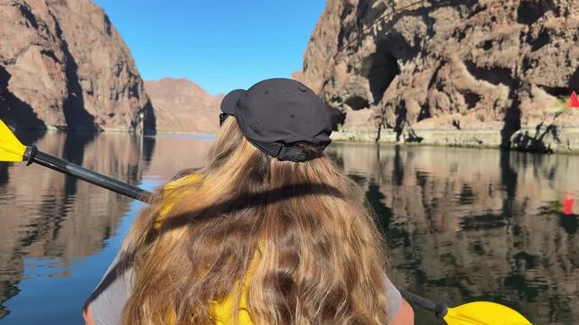 Kayaking through a rocky Colorado River canyon, a young paddler moves across calm water with steep desert cliffs reflected in the clear surface