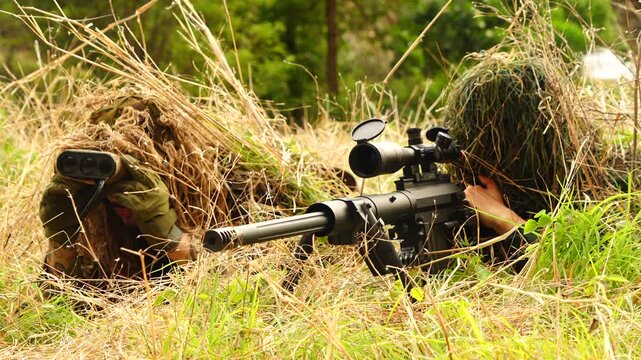  Two soldiers in camouflage uniforms stand in a field, with one soldier randomly aiming his rifle and another providing coordinates to a spotter to monitor the target.
