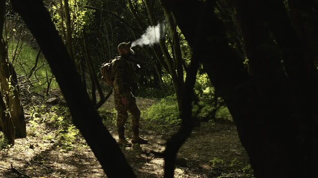 Camouflaged soldier in the middle of a forest on a recon mission. Smoking a cigarette and holding a AR-15 carbine rifle with a red collimator and a magnifier