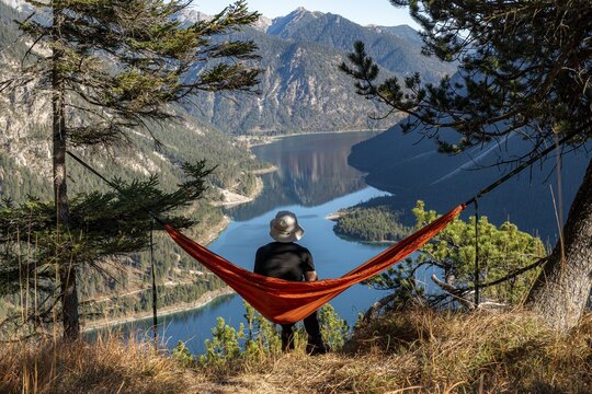 Young man sitting in red hammock with panoramic view of mountains and lake, Plansee, Ammergau Alps, Reutte district, Tyrol, Austria