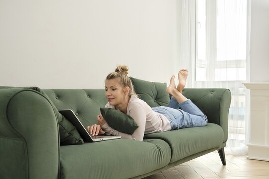 Middle-aged woman lies comfortably on a green couch, focusing on her laptop in a well-lit living room. She appears relaxed, with her feet kicked up and a pillow supporting her arms
