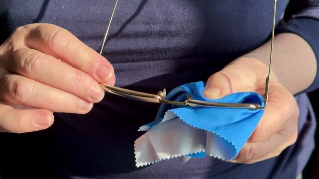 Closeup hands cleaning eyeglasses with satin cloth, woman gently polishes gold frames and lenses, sunlight highlights microfiber