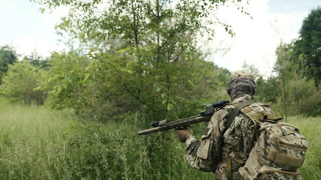 Camouflaged soldier in the middle of a summer meadow on a recon mission. Walking and carrying a AR-15 carbine rifle with a red laser collimator and a magnifier