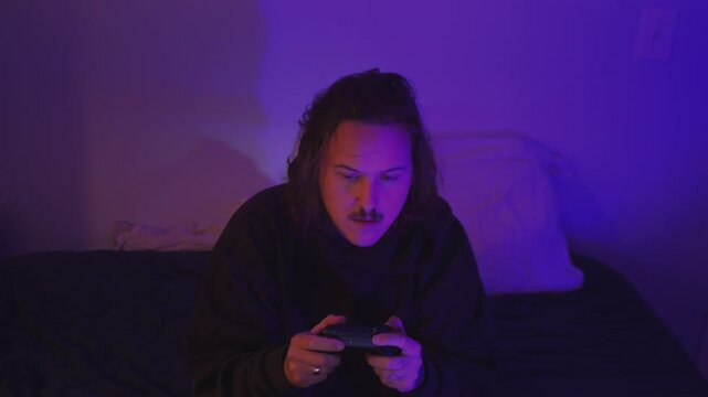 Gaming culture, technology, streaming, and home leisure. Young man plays video games with a wireless controller in a cozy bedroom illuminated by purple neon lighting.