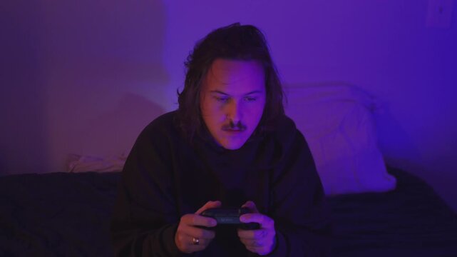 Young man plays video games with a wireless controller in a cozy bedroom illuminated by purple neon lighting.