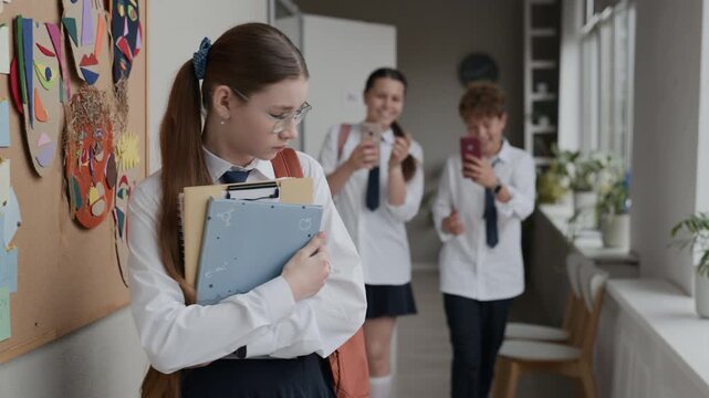 Caucasian teenage girl looking down and holding folders in school corridor while classmates mocking and filming her. Video perfect for cyberbullying awareness and school safety campaigns