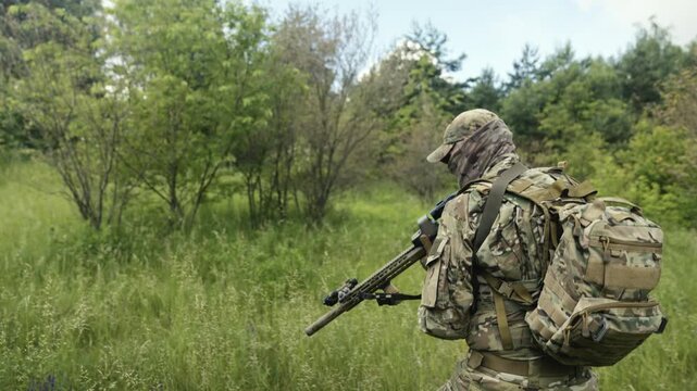 Camouflaged soldier in the middle of a field of wild grass on a mission. Walking and aiming a AR-15 carbine rifle with a red laser collimator and a magnifier