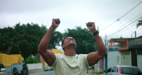 Obraz premium Man of African descent standing on urban street with arms raised, looking up in gratitude, emotional moment of relief and hope, expressive gesture of thankfulness and faith