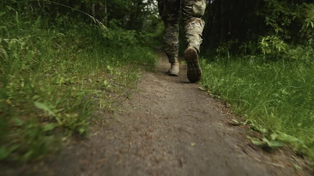 Legs of a camouflaged soldier in the middle of a footpath in a forest on a recon mission. Walking and aiming a AR-15 carbine rifle with a red laser collimator and a magnifier