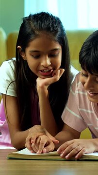 Indian small brother sister studying reading book together at home indoors, Asian young kids siblings learning, focusing on education activity in modern living room, childhood development with family