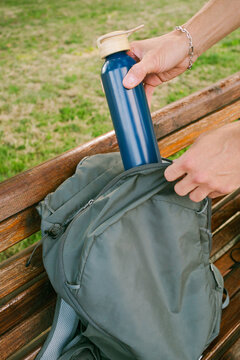 Person Puts a Water Bottle Into a Backpack on a Wooden Bench