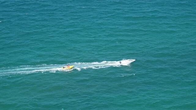 A white motorboat tows a yellow inflatable carrying people across the clear blue ocean, creating a foamy wake. This exciting watersport takes place on a bright day