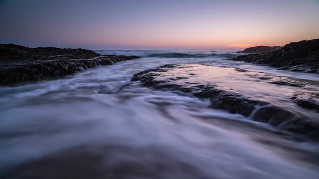 Slow shutter speed ocean waves flowing over dark rocks on a sandy beach during sunset, creating a soft misty water effect with a clear horizon line and colorful dusk sky.