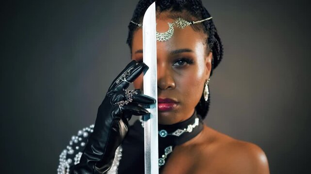 closeup portrait african fantasy woman warrior queen holding sword in hands strokes blade, sexy looks at camera. Beauty face black dress girl princess tiara moon, dark gray studio. vertical video.