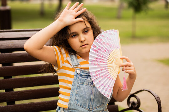 Cute little girl kid child is sitting on a park bench, fanning herself with a decorative fan to cool off.