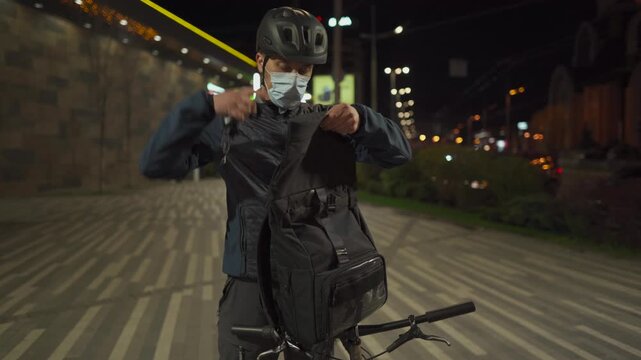 Night urban scene of a male cyclist wearing a helmet and face mask opening a rolltop backpack and storing a bicycle chain lock. City lights and modern transportation lifestyle in a big city.