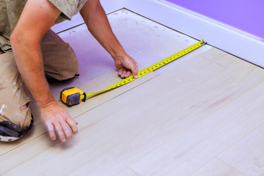 Carpenter worker uses tape measure to measure wooden floorboards in room with purple walls.