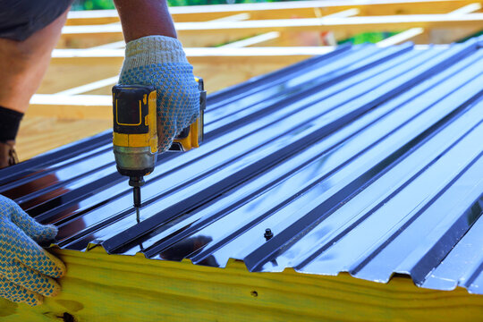 Worker in gloves drills metal sheets onto wooden beams frame at construction site