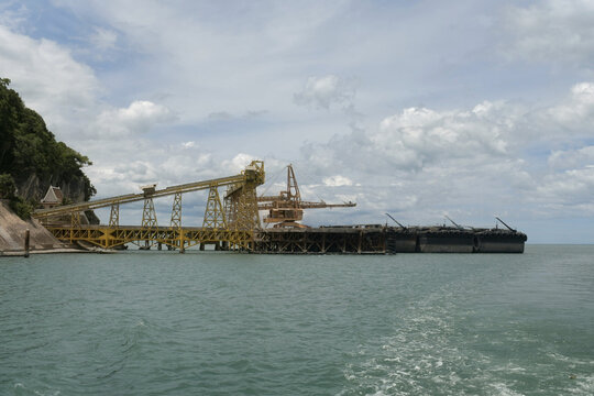 industrial belt conveyor system terminal loading mineral materials into large cargo barge vessel at khanom coastline jetty facility.