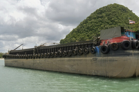 large industrial cargo barge transport vessel moored at khanom shoreline terminal facility with lush green tropical mountain background