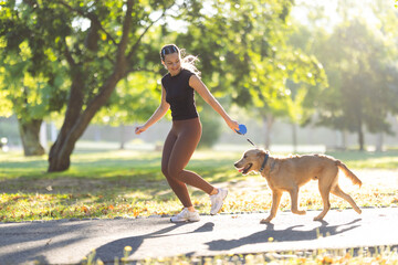 Obraz premium Young woman walking a golden retriever dog on a sunny day in a park, wearing athletic clothing and enjoying outdoor exercise along a paved path