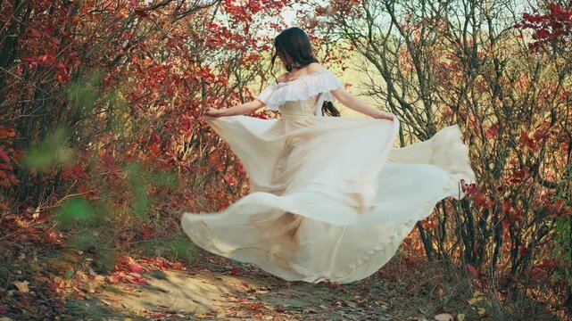 Fantasy happy woman enjoy nature dancing in forest. Princess girl beauty smiling face. White long vintage dress waving, dark hair fly in motion Lady dancer. Autumn nature red orange leaves trees park