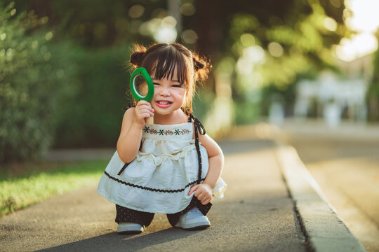 Cute little Asian girl holding magnifying glass to her eye and smiling while crouching outdoors, happy curious child playing in green park for early childhood development, science learning, and parent