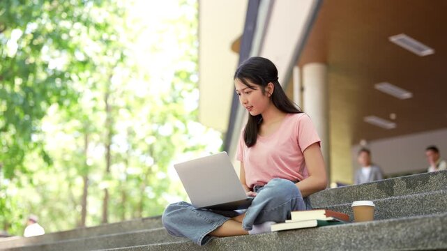 Smiling young asian young woman in university sitting at building stair working on laptop computer working or studying.