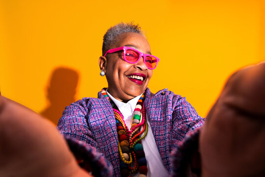 Senior woman smiling taking a colorful selfie - Old hispanic grandmother wearing fashionable modern clothing on colorful background, concepts about elderly, aging, diversity 