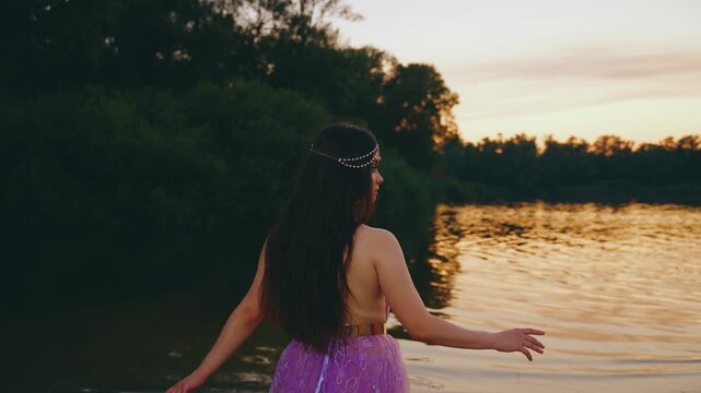 Fantasy woman elf queen lady sitting on river bank, purple long dress back rear view. fairy girl creative costume. Summer dark nature water lake river sky red sunset divine magic sun light rays art 4k