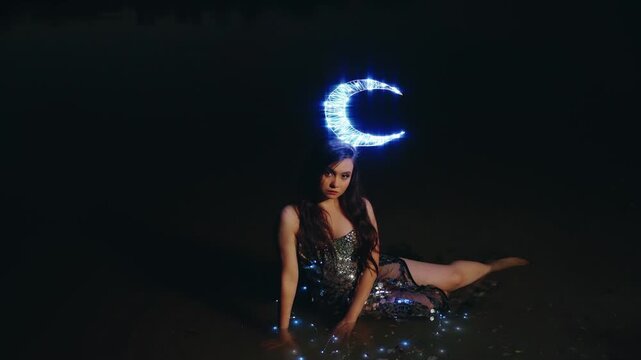 Fairy girl fashion model sitting in water lake river. Fantasy woman queen of night lady moon, crown glows on head, black dress cape shining stars costume back rear view. Summer dark nature dusk sky 4k