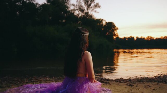 Fantasy woman elf queen lady sitting on river bank, purple long dress back rear view. fairy girl creative costume. Summer dark nature water lake river sky red sunset divine magic sun light rays art 4k
