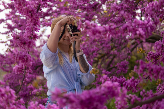 Young woman smiling while capturing photos with a vintage camera, embracing springtime amidst a profusion of beautiful purple glicinia flowers in bloom