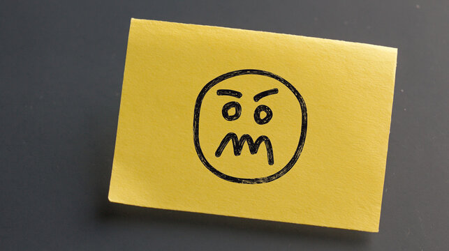 Yellow sticky note with angry face drawing on black background. Sticky note with hand-drawn emoji