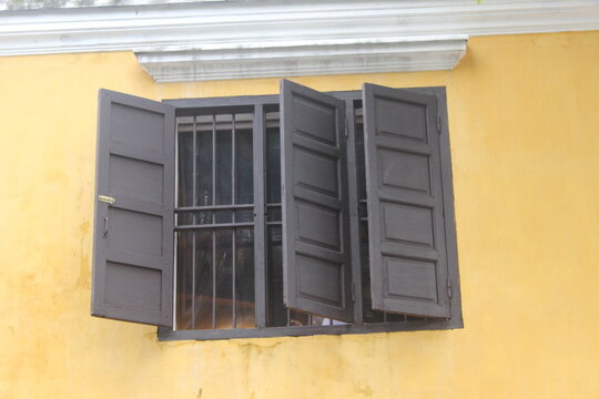 Old brown wooden window frame with open shutters on yellow plastered exterior wall.