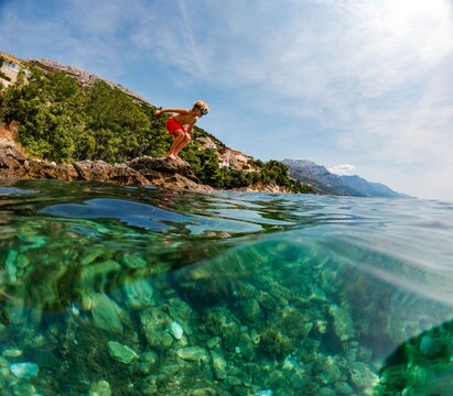 Young boy jumping from coastal rocks into Adriatic Sea on family holiday.