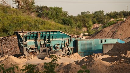 Słoneczny kadr przedstawiający mobilną, niebieską kruszarkę gąsienicową pracującą przy hałdach piasku w kopalni odkrywkowej lub na placu budowy. © tomo81