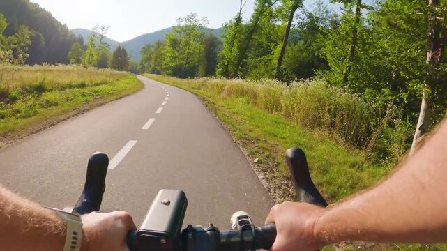 POV of cyclist riding on asphalt bike path at sunset in summer. Slovenia. First person view of male hands on bicycle handlebar cycling on road in green fields, forest. Active sport outdoor lifestyle