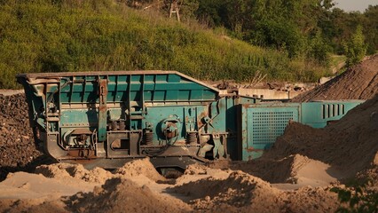 Słoneczny kadr przedstawiający mobilną, niebieską kruszarkę gąsienicową pracującą przy hałdach piasku w kopalni odkrywkowej lub na placu budowy. © tomo81