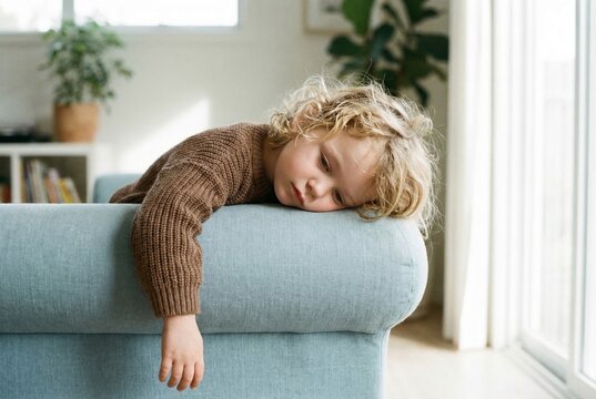 Sad and tired little boy lying down over the armrest of a blue sofa at home