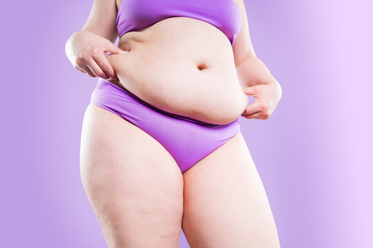 Overweight woman pinching belly fat in underwear on purple background. Obesity, weight loss, diet and body positivity concept