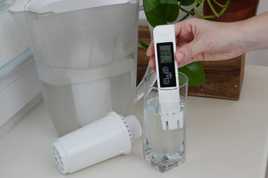 Hand measuring filtered drinking water with a digital TDS meter beside a household filtration pitcher and replacement cartridge. Concept of water purification, clean drinking water, water quality test
