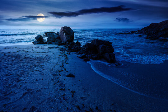 huge stones on the sandy beach at night. mysterious holiday season background at the sea. full moon over clouds on the dark sky above horizon. view of waves washing the shore
